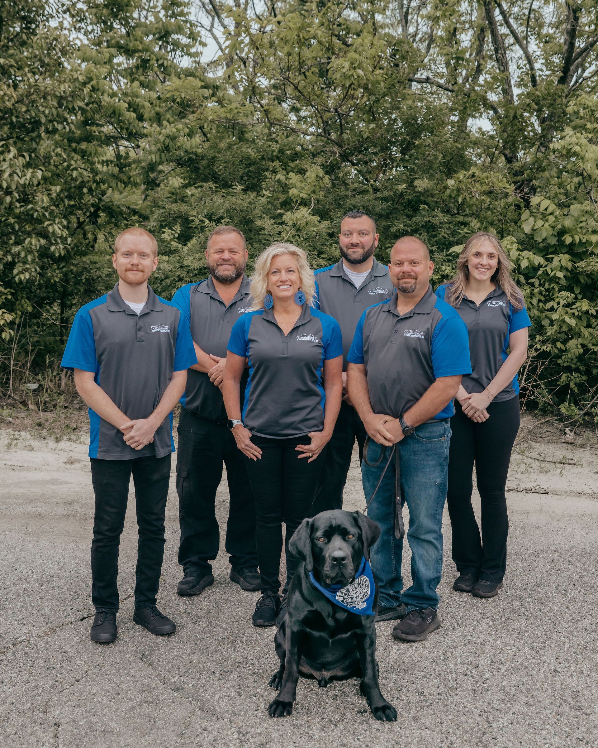 Five people in blue uniforms stand indoors with a black service dog in front of a white wall.