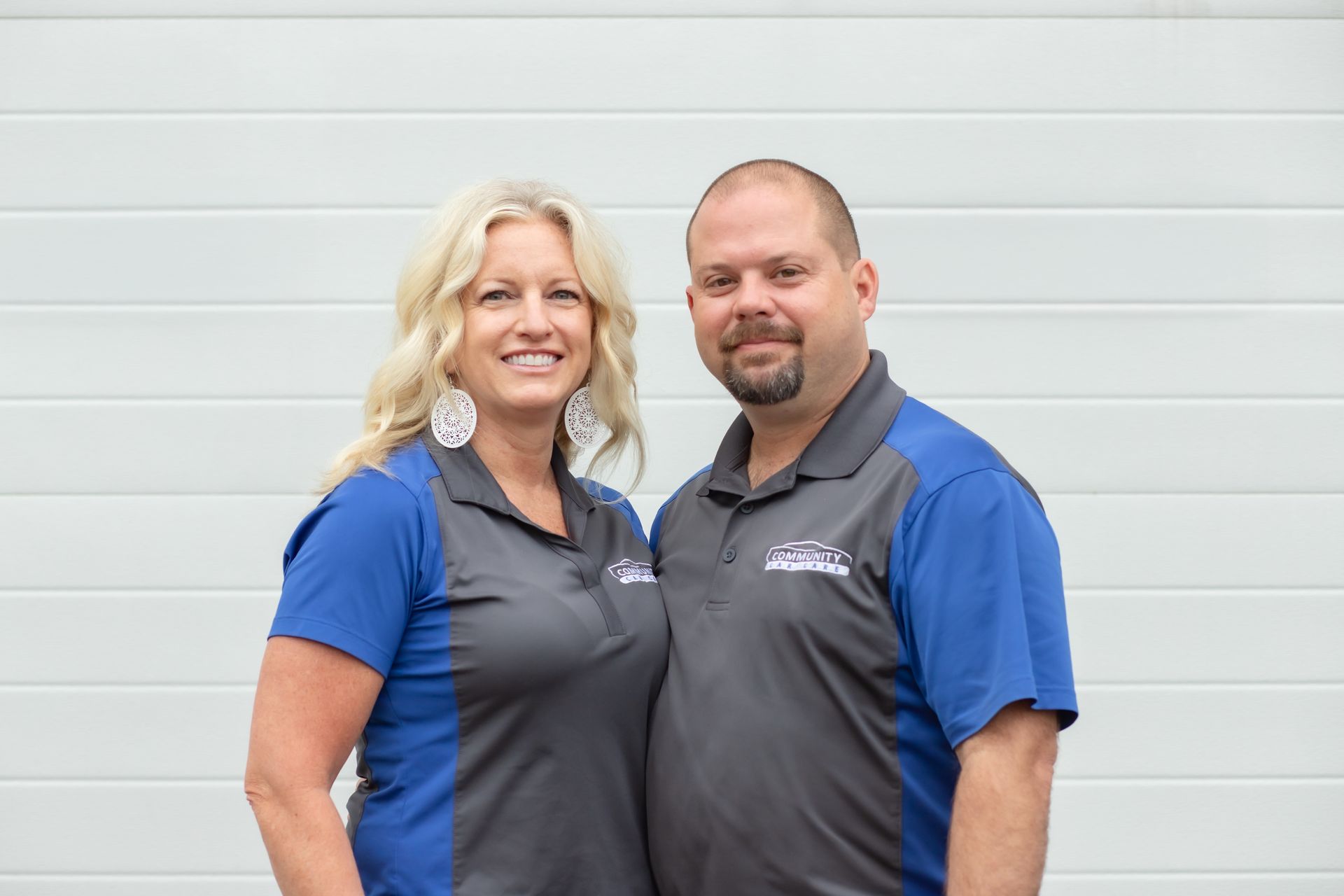 Two smiling employees in blue and gray shirts standing against a white wall
