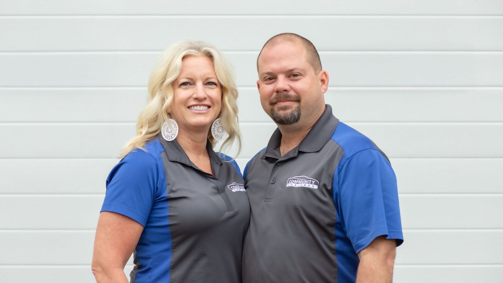 Two coworkers in blue shirts and gray vests standing and smiling in front of a white wall