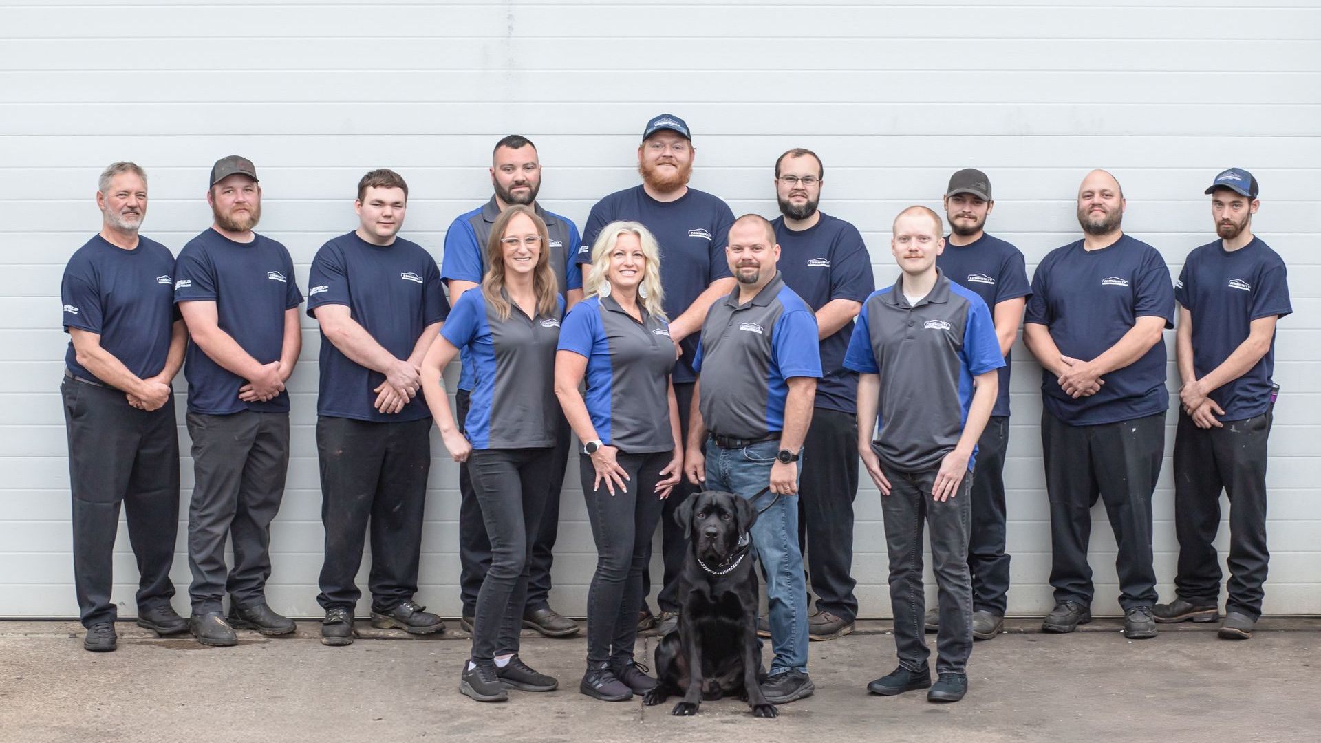Uniformed team posing with a black police dog in front of a white garage door.