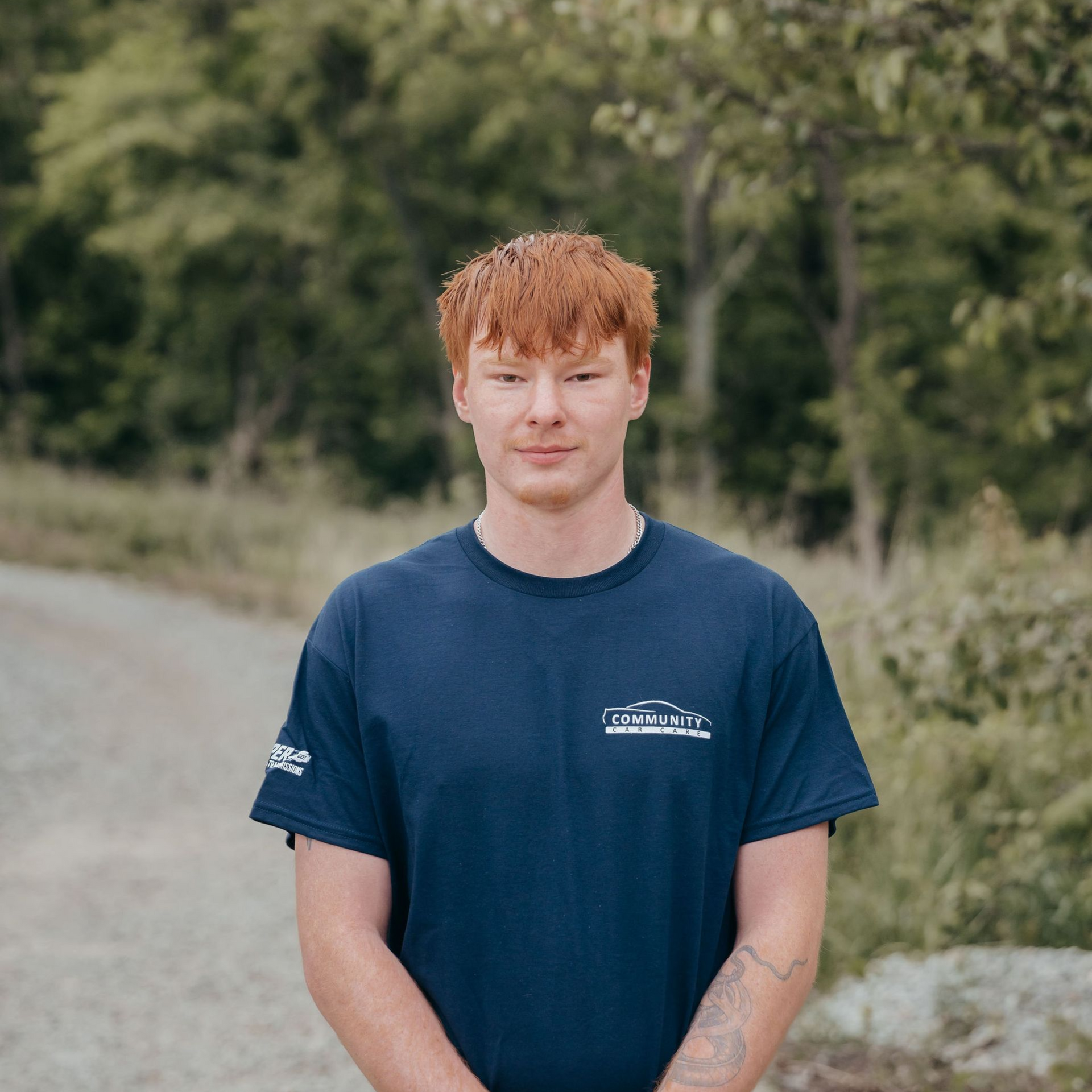 Person with curly red hair wearing a navy T-shirt stands outdoors on a road with trees behind them.