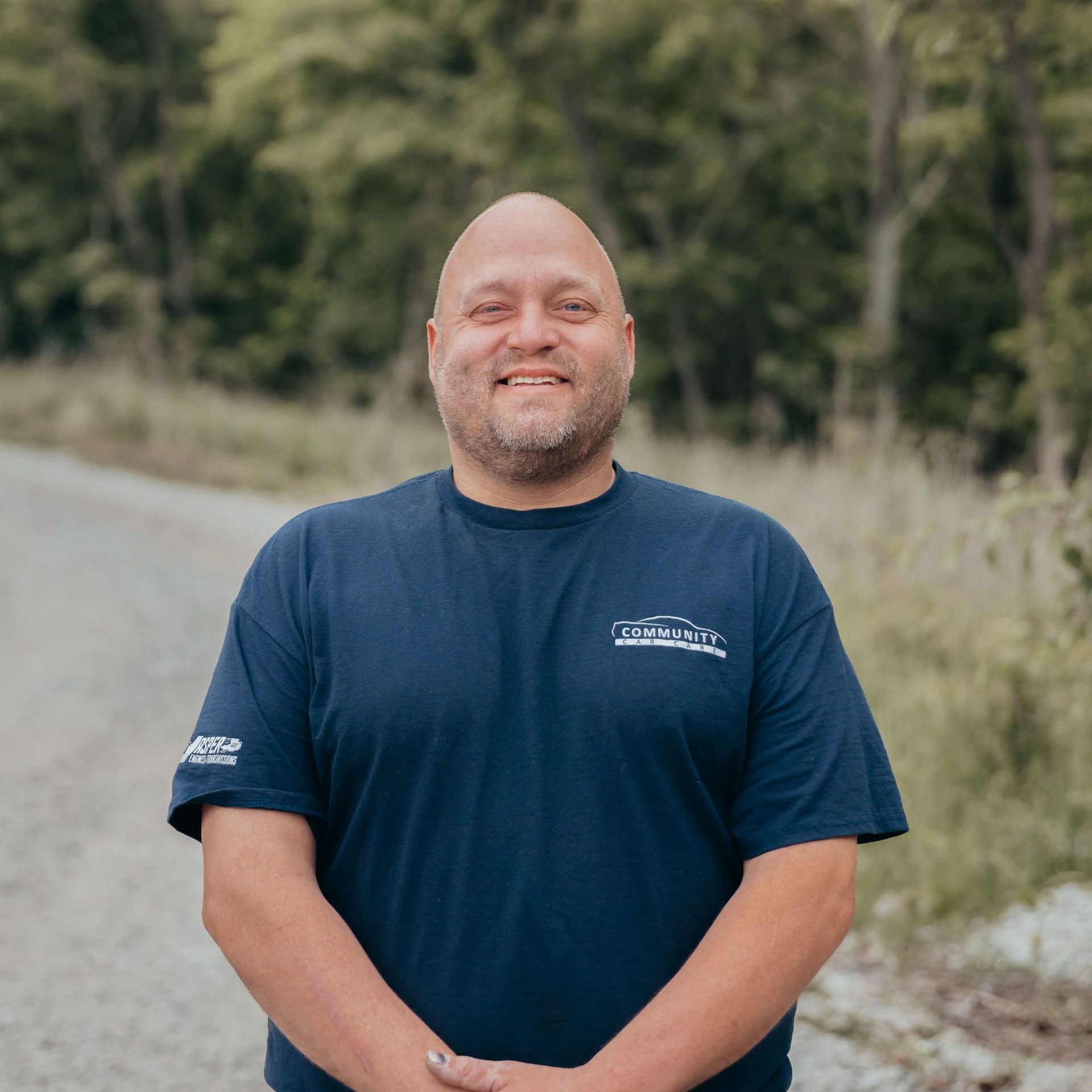 Smiling bald man in a navy T-shirt standing outdoors on a dirt road with trees behind him