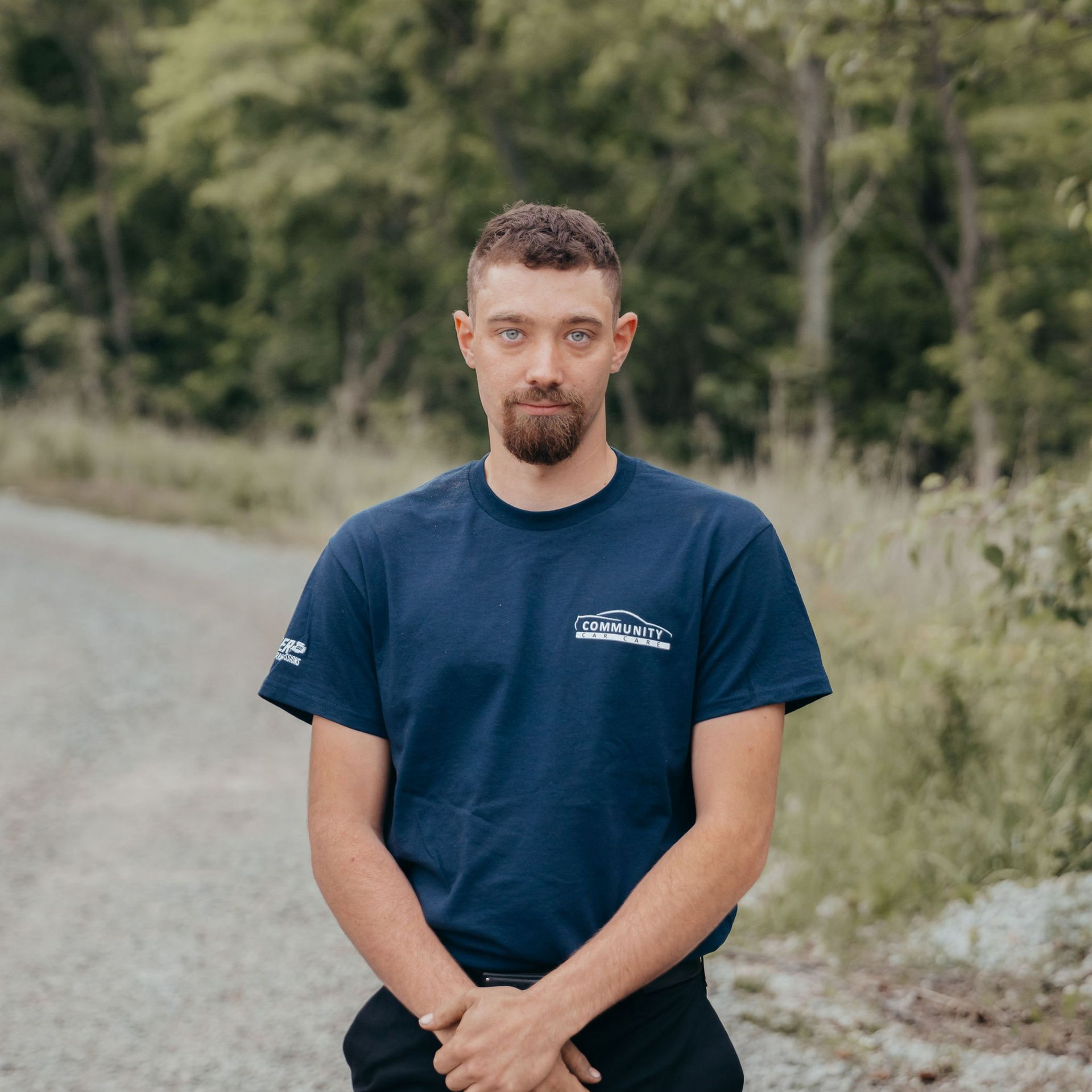 Man standing on a dirt road in a navy T-shirt, with trees and grass in the background.