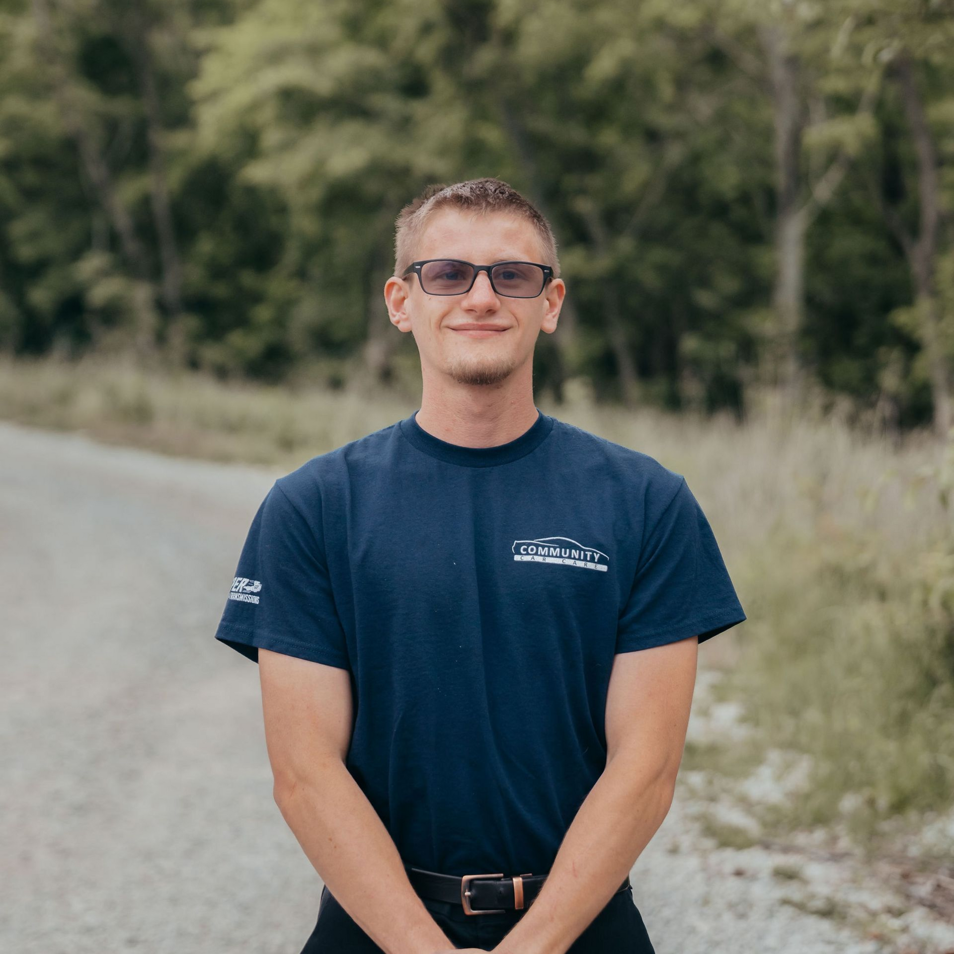 Person standing outdoors on a dirt road, wearing sunglasses and a navy T-shirt, with trees in the background.