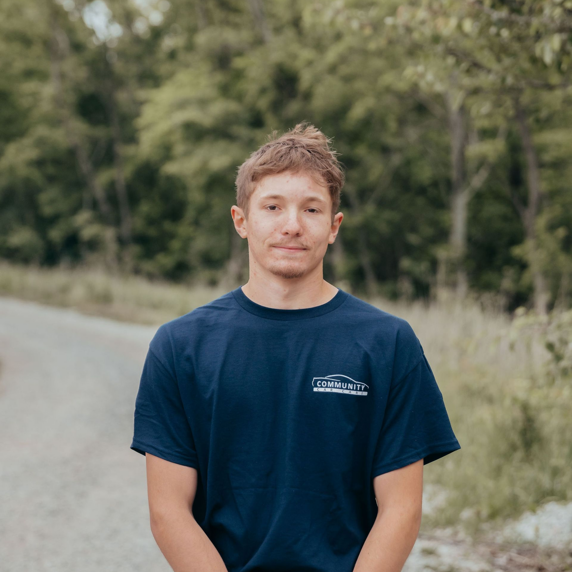 Person standing on a rural road with trees in the background, wearing a navy T-shirt and slight smile