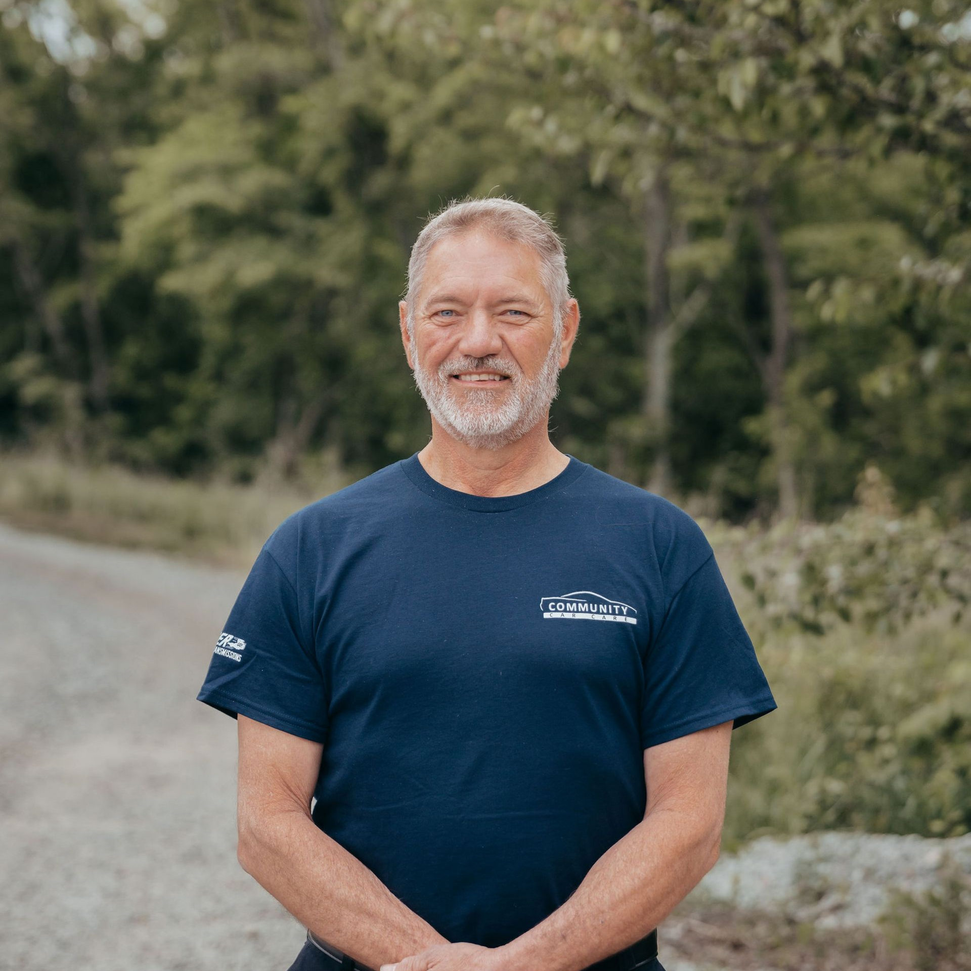 Smiling man in a navy T-shirt standing outdoors on a gravel path with trees behind him