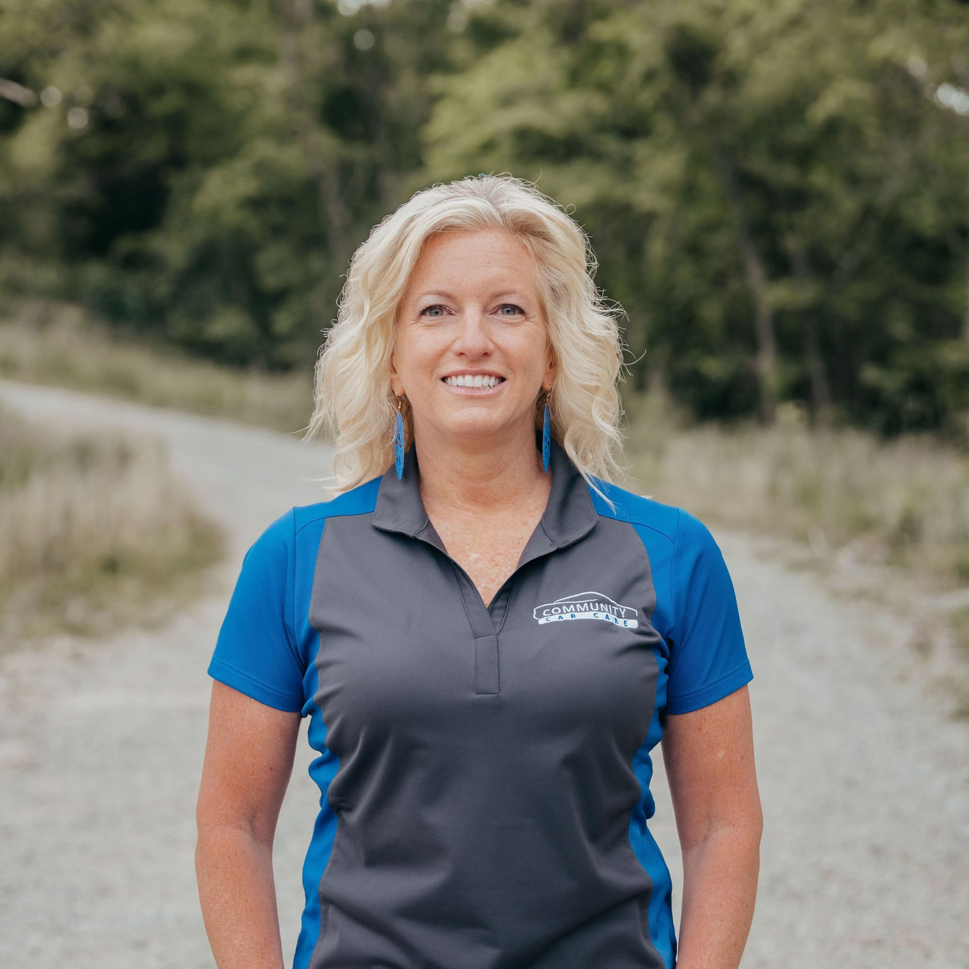 Smiling woman in blue and gray polo standing outdoors on a sandy path