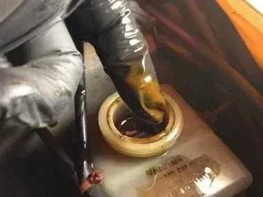 Gloved hand reaching into a car's brake fluid reservoir filled with dark, dirty-looking fluid.