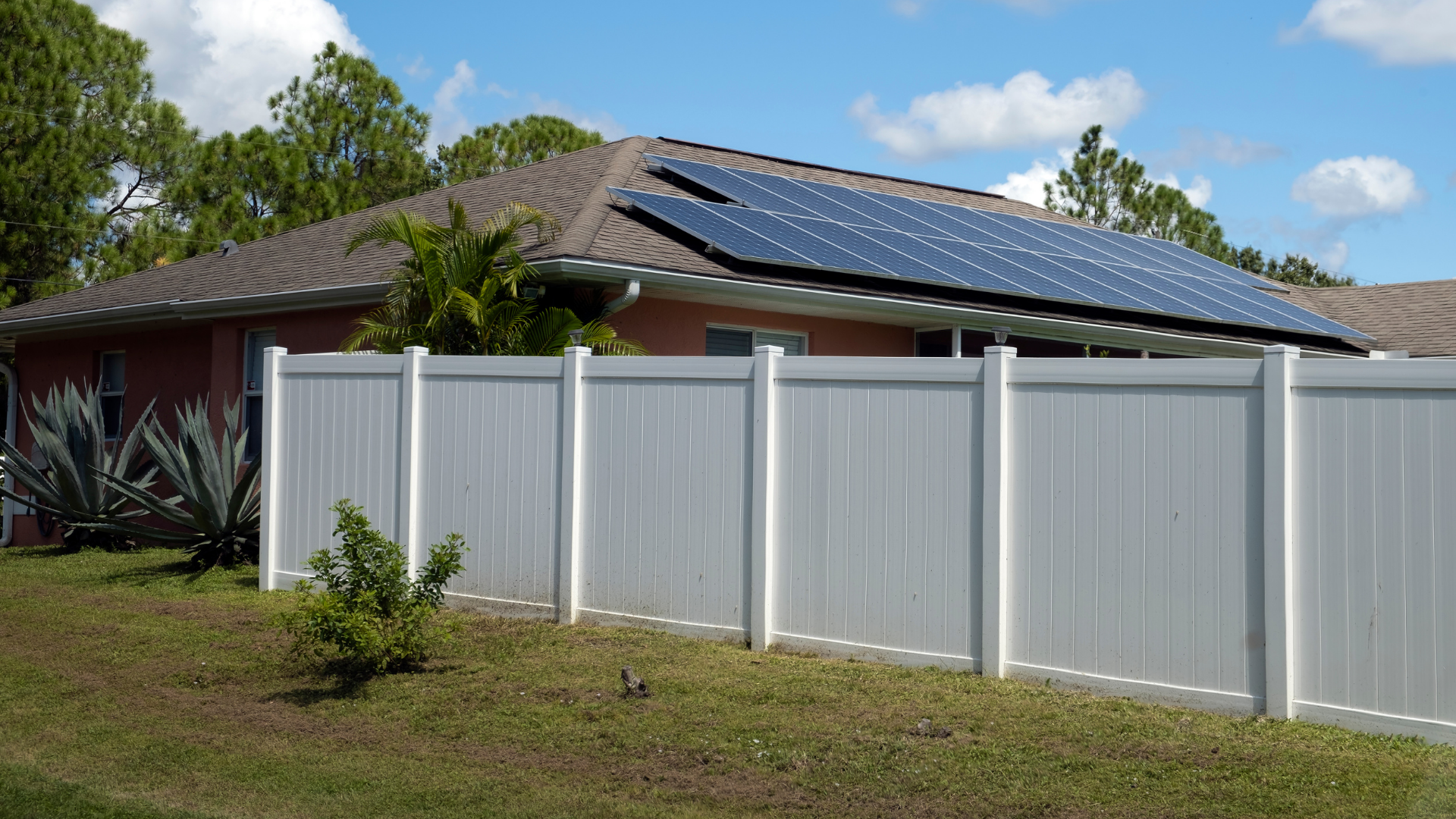 Suburban house with solar panels behind a white fence under a blue sky