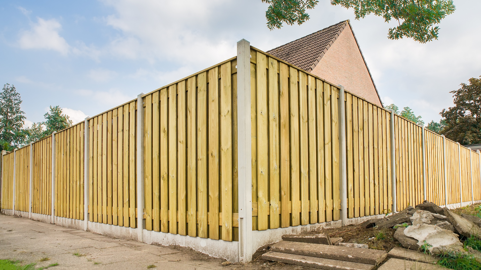 Yellow wooden fence around a house with a tiled roof and stone steps on a sandy lot.