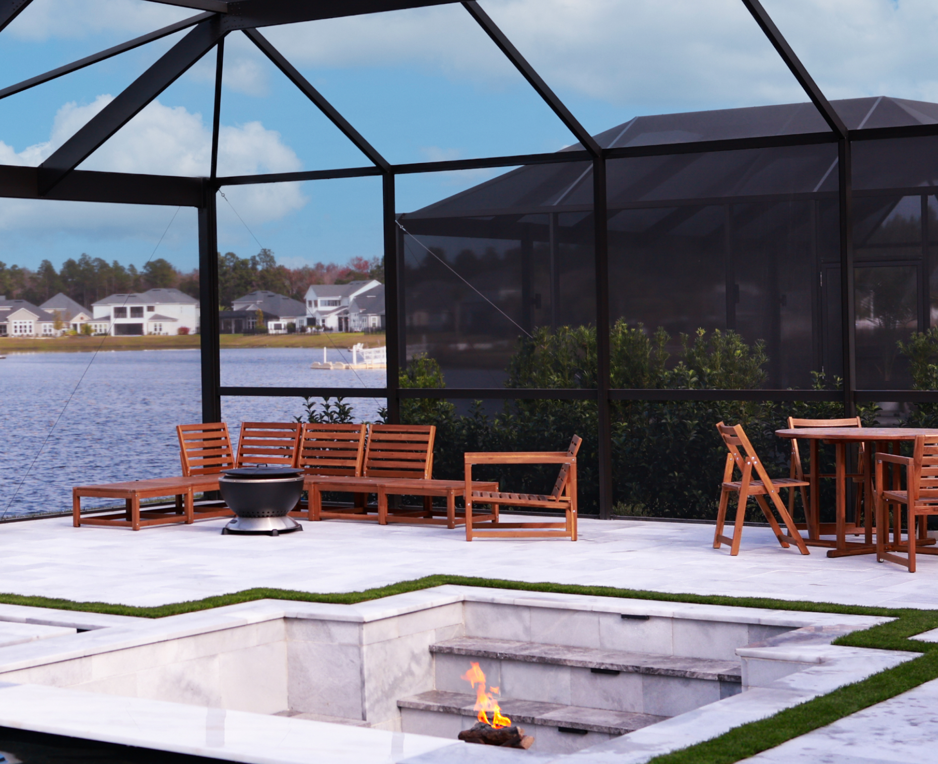 A modern outdoor sunken seating area with a central fire pit on a patio, overlooking a lake within a screened enclosure.