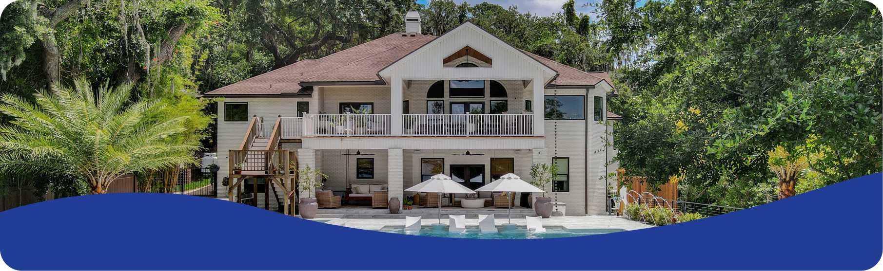Two-story cream-colored house with a balcony, patio, and swimming pool surrounded by lush green trees.