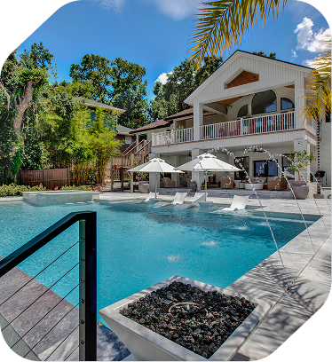 A sunlit swimming pool with white umbrellas and lounge chairs in front of a modern two-story home with a stone patio.