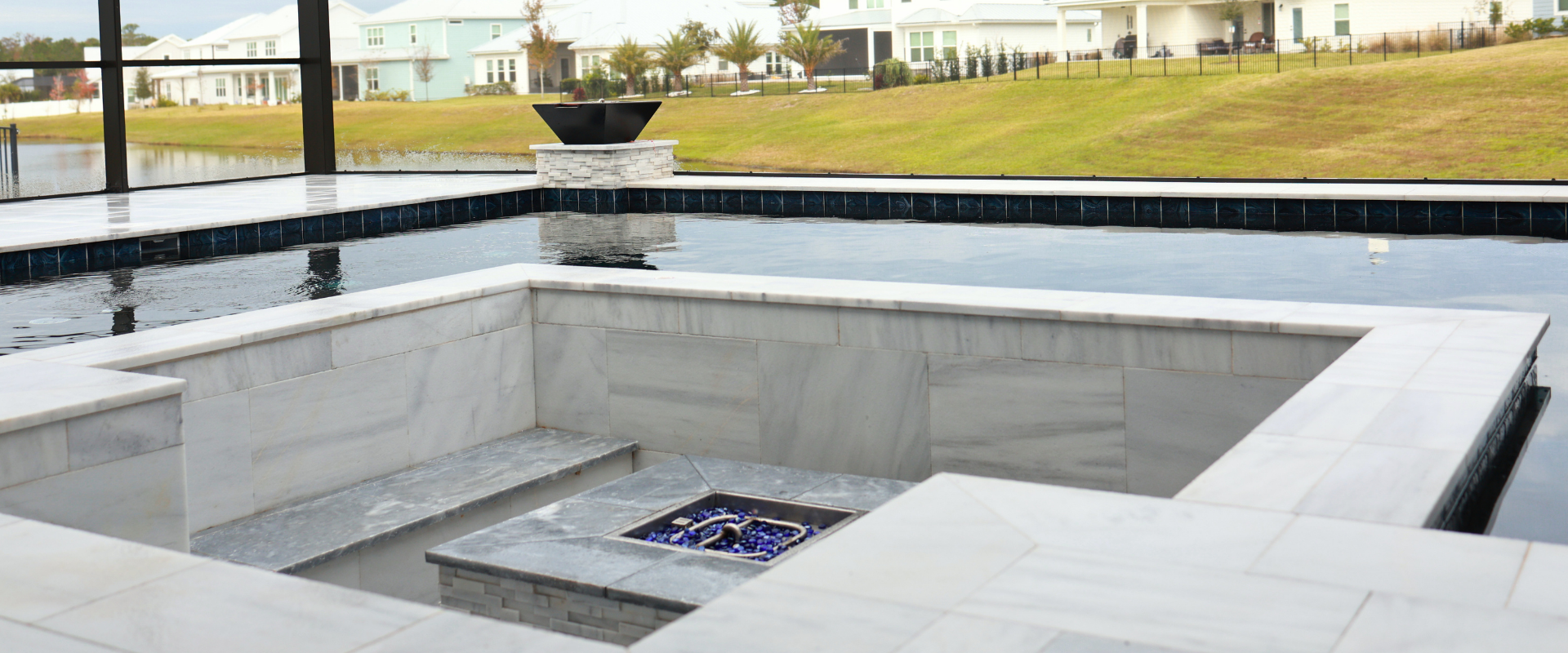 A sunken fire pit area with light stone walls and blue glass fire rocks, centered inside a modern swimming pool.