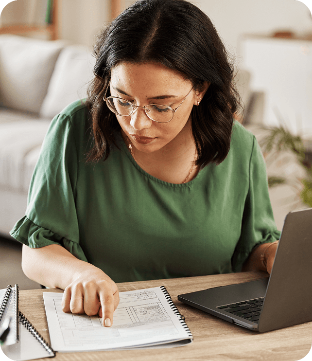 Woman in green shirt reviewing notes beside a laptop at a desk