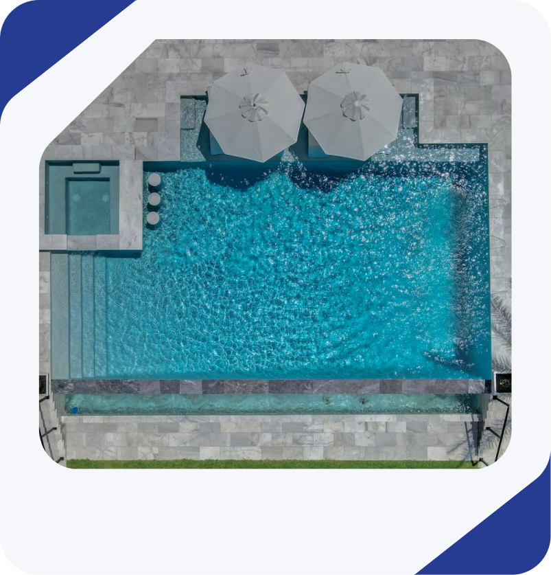 Overhead view of a rectangular turquoise pool with a jacuzzi, umbrellas, and stone patio.