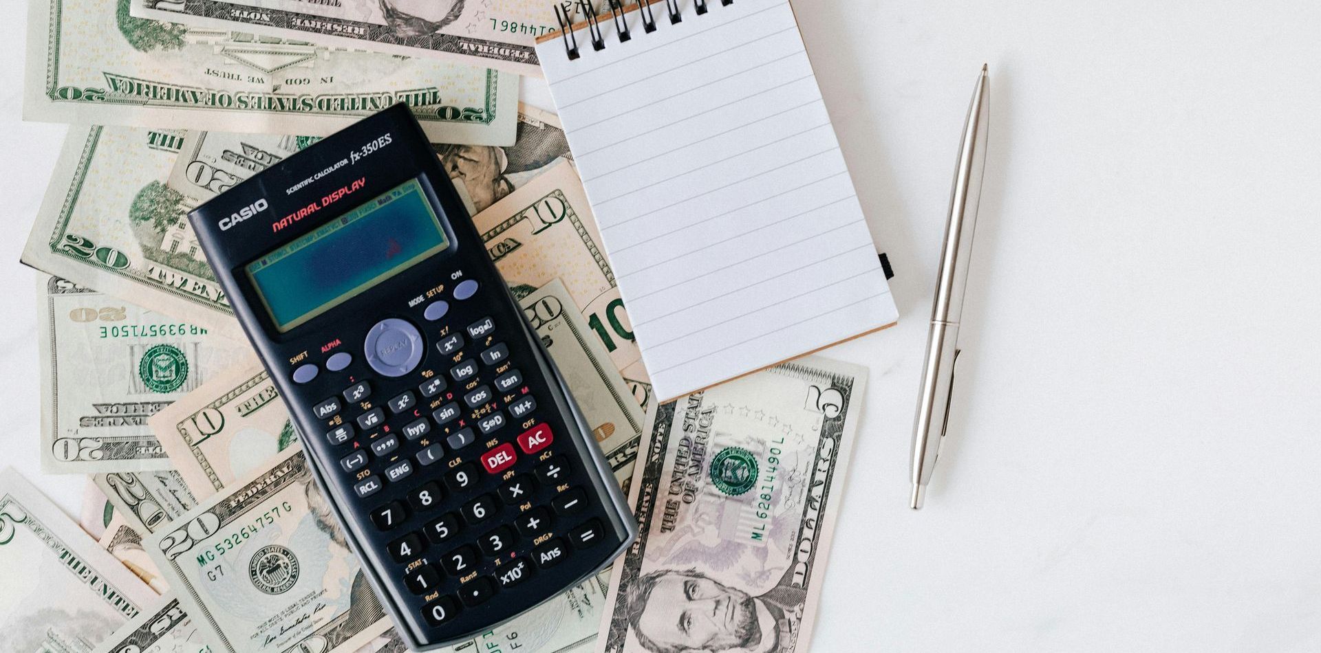 A calculator is sitting on top of a pile of money next to a notebook and pen.