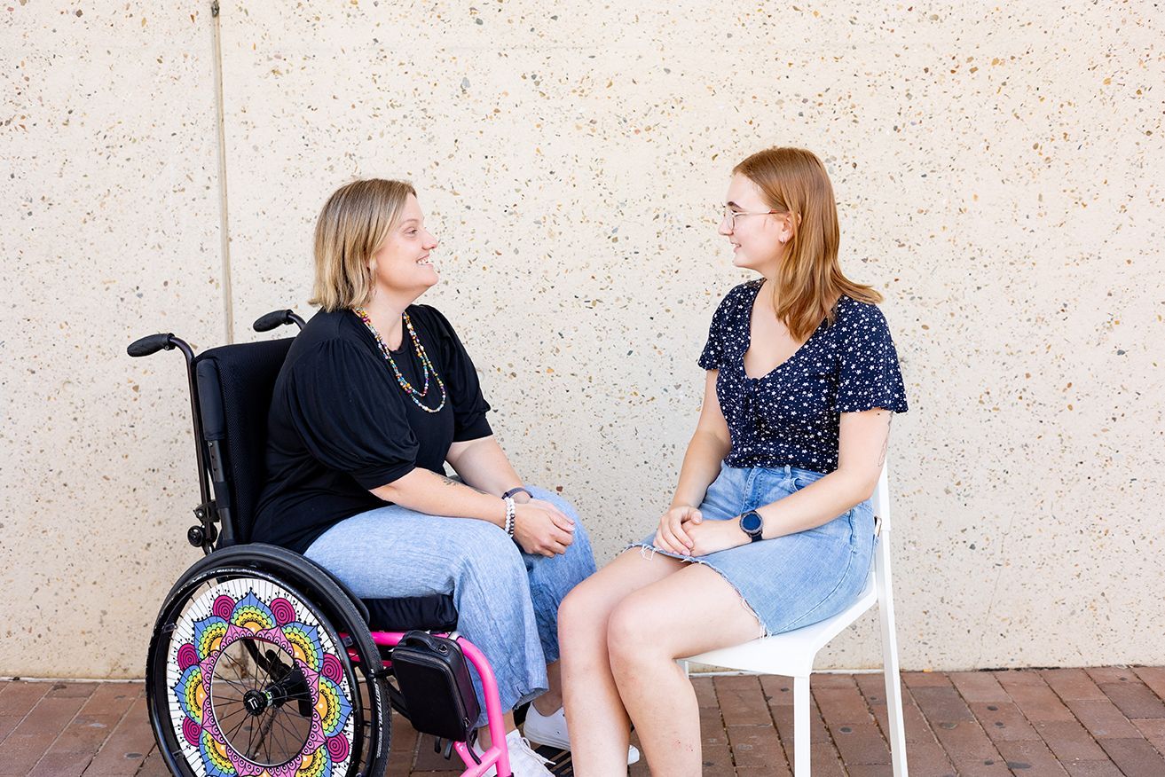 two people sitting outside with a concrete wall behind them. One is a wheelchair user.