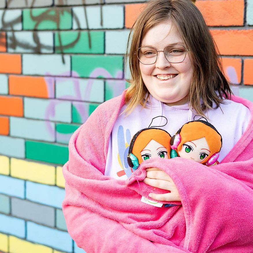 An individual is standing Infront of brick wall smiling. The individual is holding two soft dolls while wrapped in a blanket.