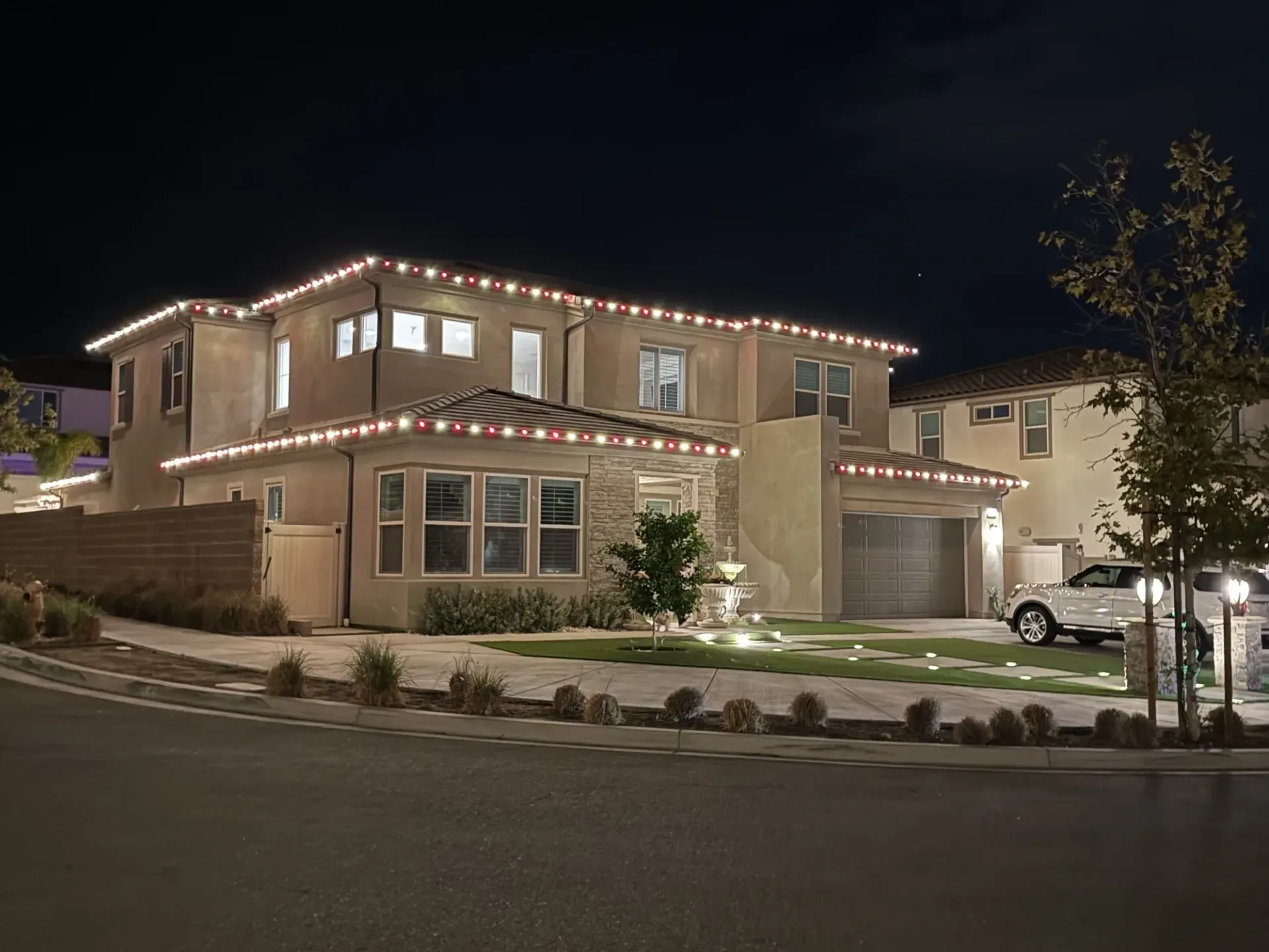 Two-story suburban house at night, outlined with white holiday lights on a corner lot.