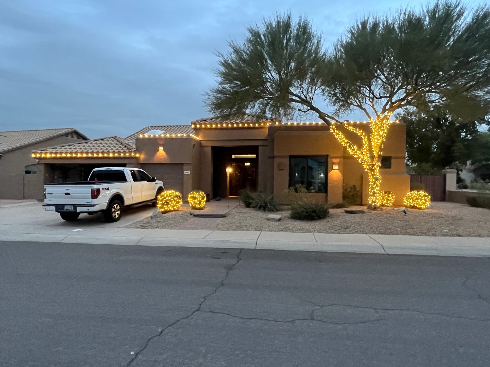 Single-story house at dusk with warm string lights, a white SUV in the driveway, and a lit tree in front yard
