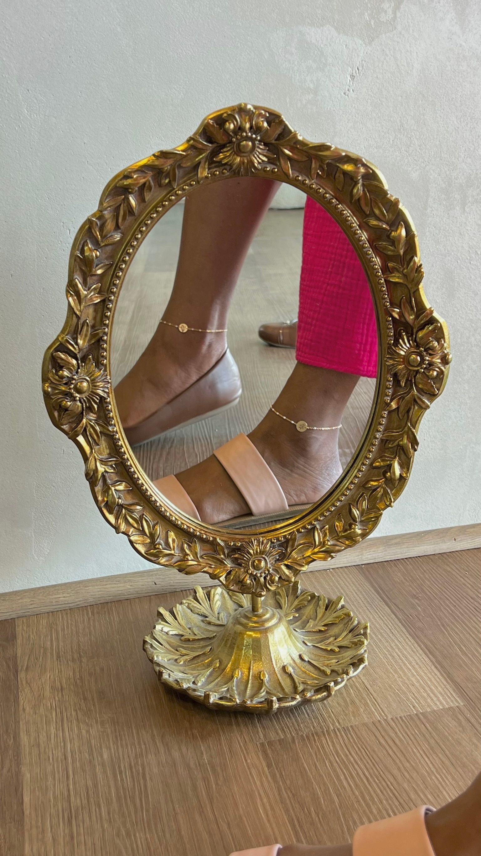 Gold decorative tabletop mirror reflecting a person’s feet in sandals and bright pink pants