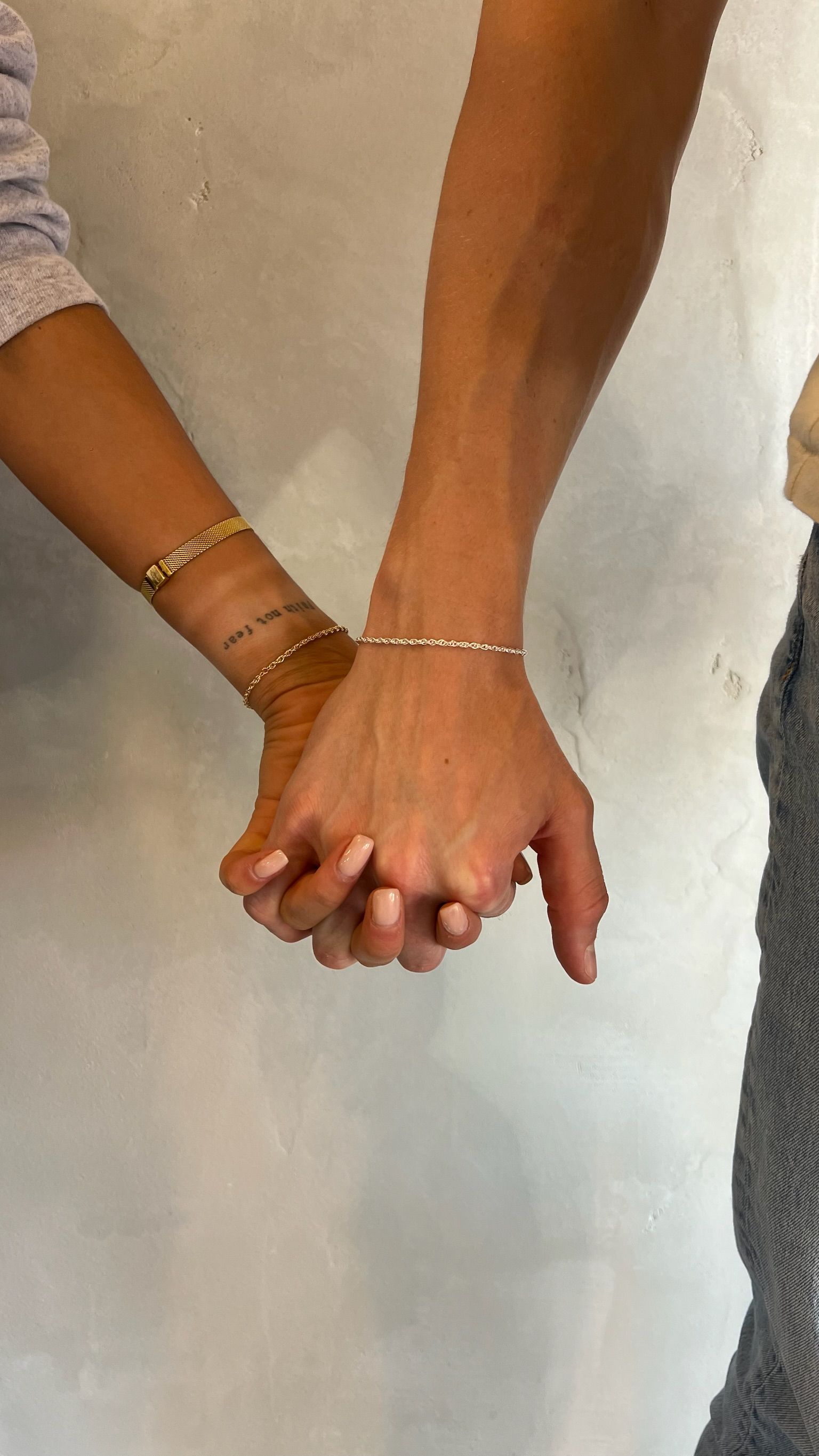 Two people holding hands, one wearing a bracelet, against a plain wall.