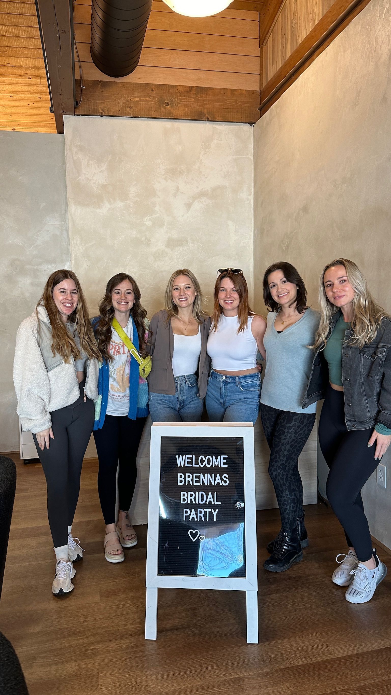 Six women pose indoors beside a signboard in a rustic room with wood beams.