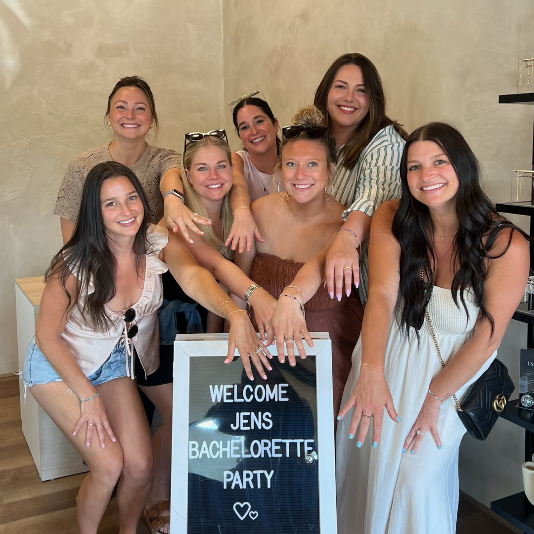 Six women posing indoors around a sign that reads “Welcome Jens Bachelorette Party”