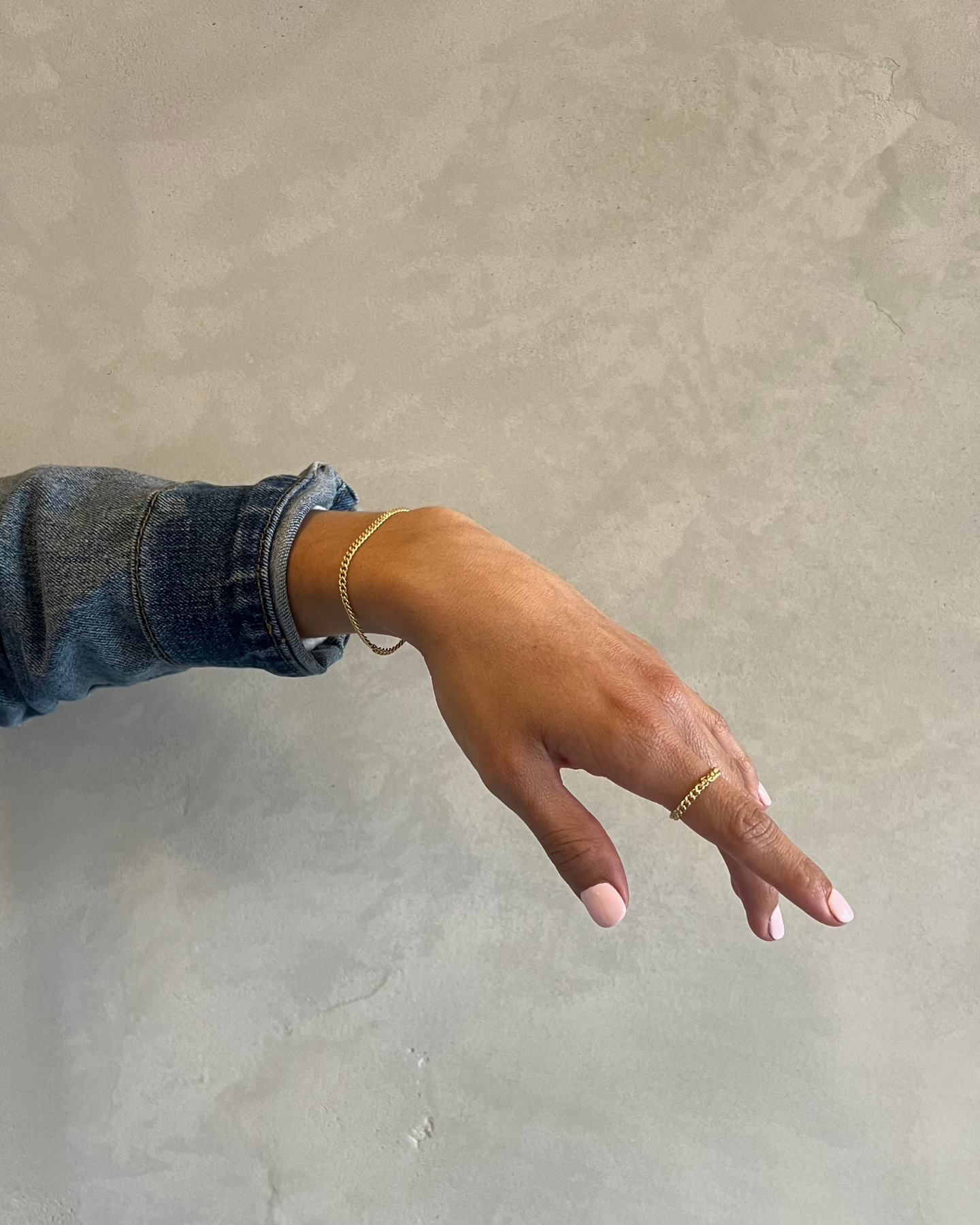 Hand with gold jewelry and white nails, wearing a denim sleeve, against a gray background