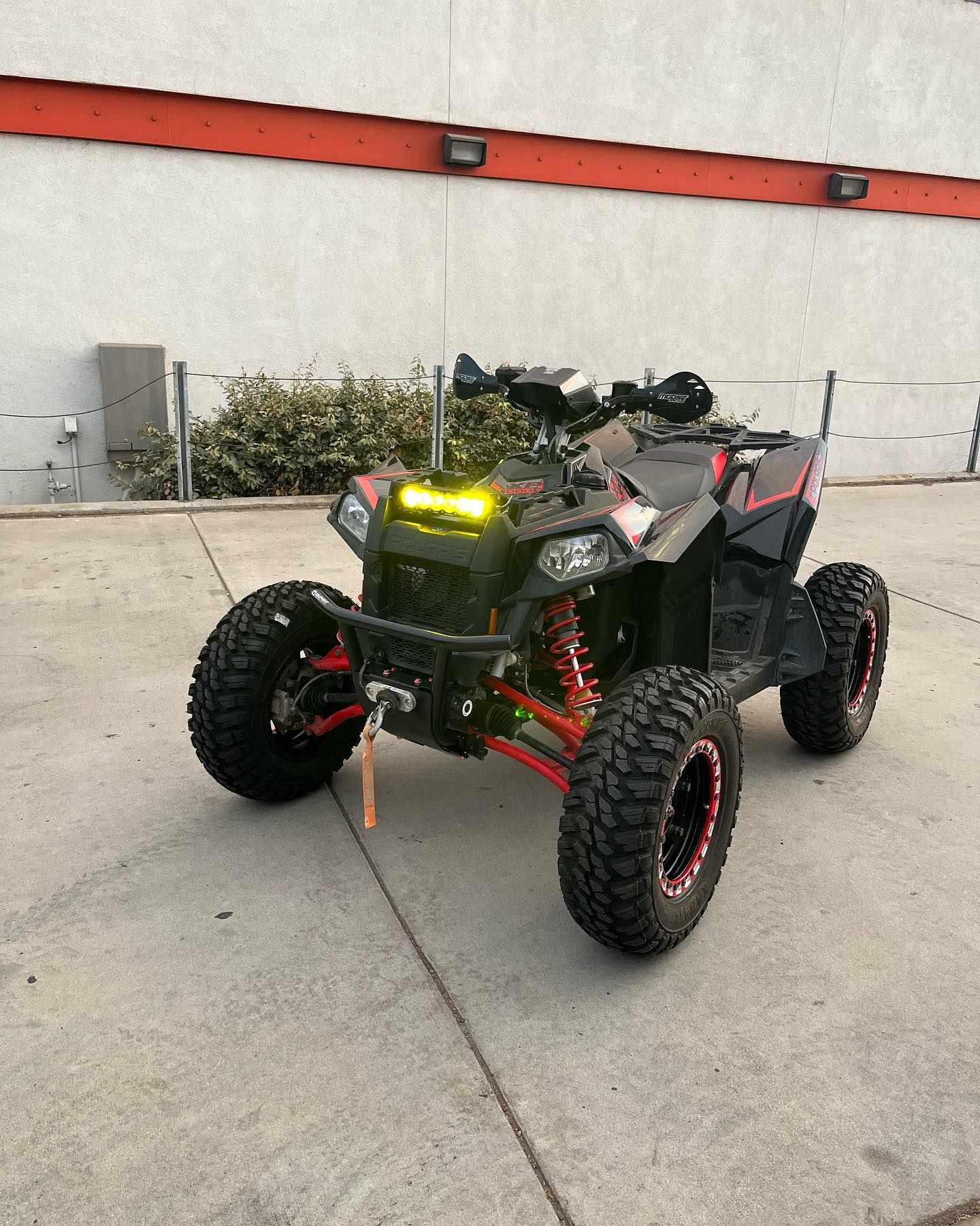 Red and black off-road ATV parked on a concrete surface with headlights on near a white wall.