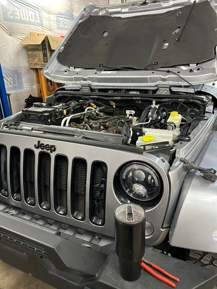 Silver Jeep with hood open, showing engine. Black grill, tools on bumper. Workshop setting.