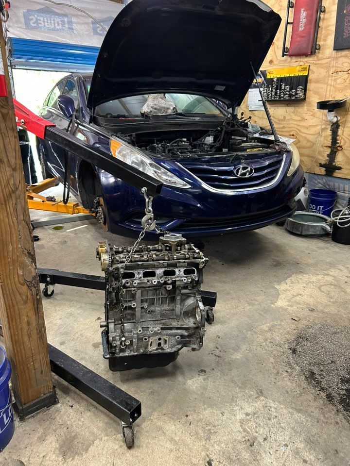 A blue car with its hood open, engine being lifted out by a crane in a garage.