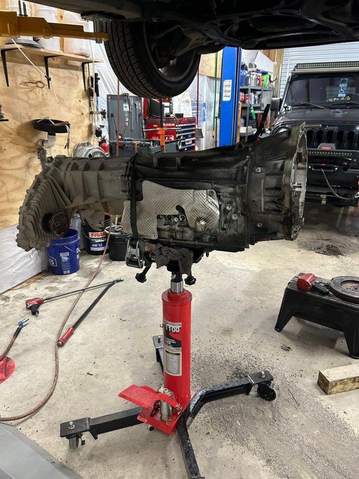 Transmission on a hydraulic jack beneath a vehicle in a garage setting. Tools and equipment visible.