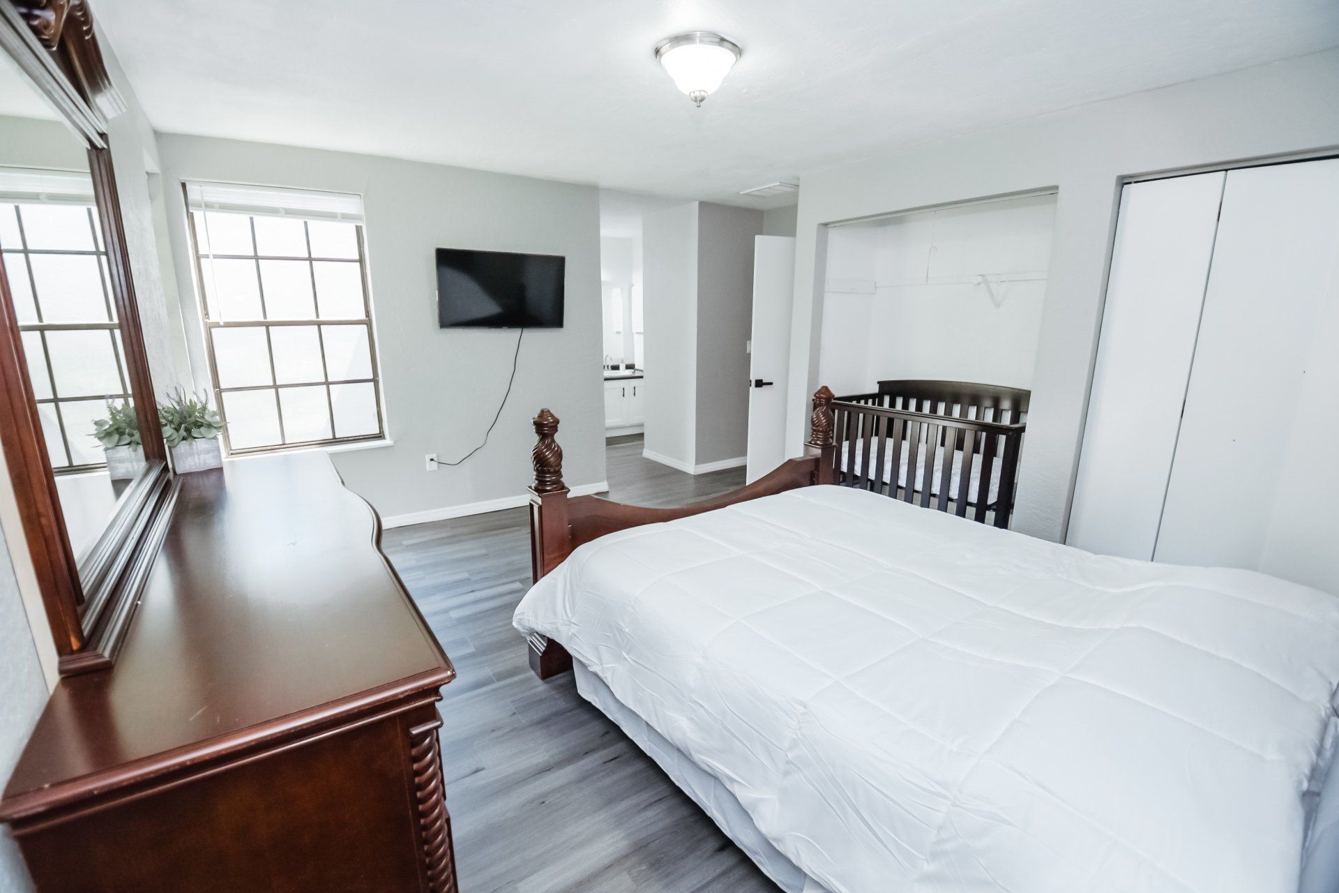 Bedroom with bed, dresser, TV, crib, and window. Gray walls and wooden floor.