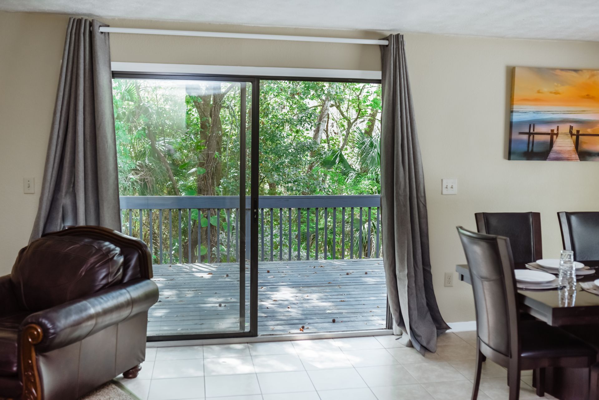 Sliding glass door opens to deck with lush greenery; gray curtains frame the view. Interior features furniture.