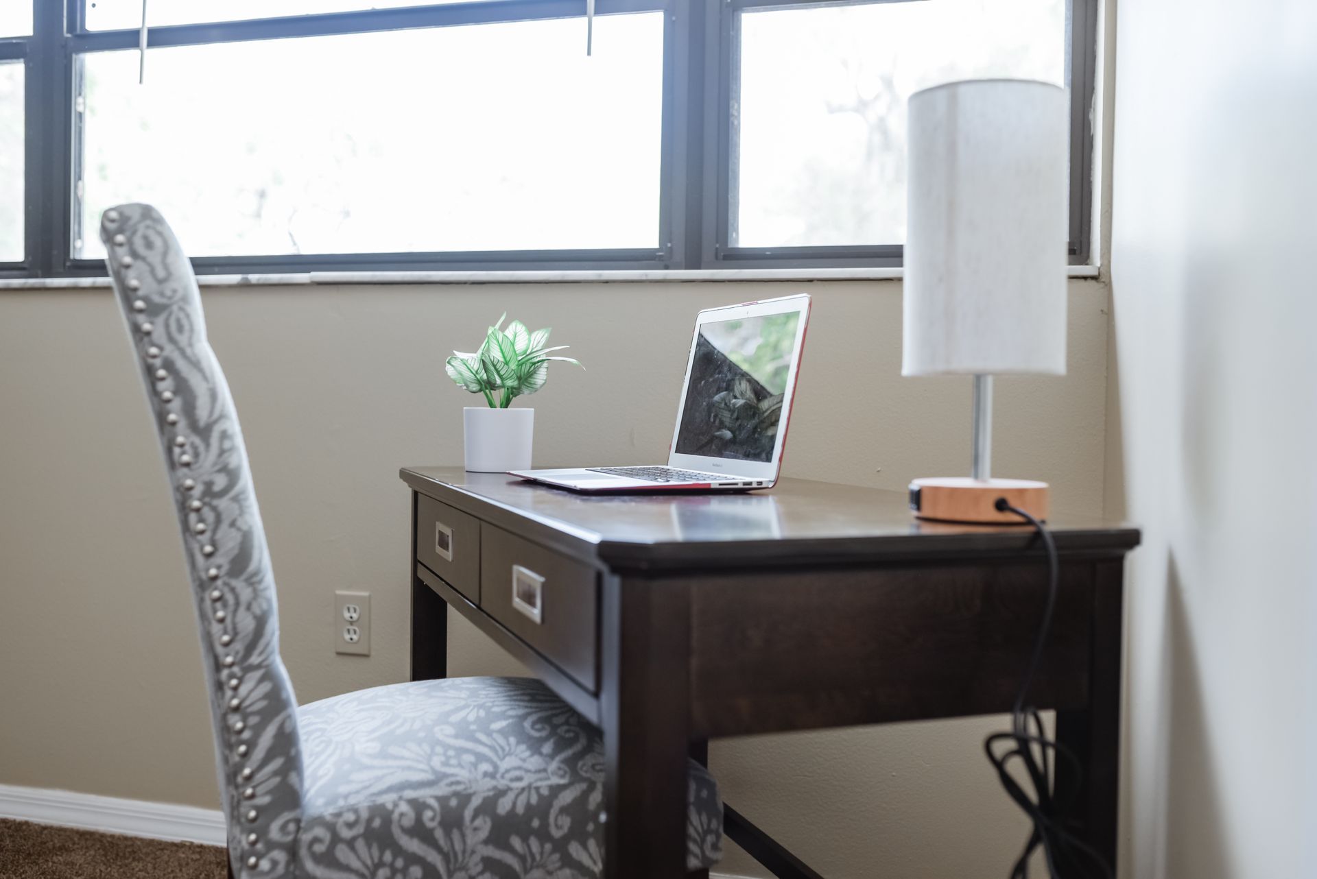 Desk with laptop, lamp, and chair by window. Artificial plant sits on desk.