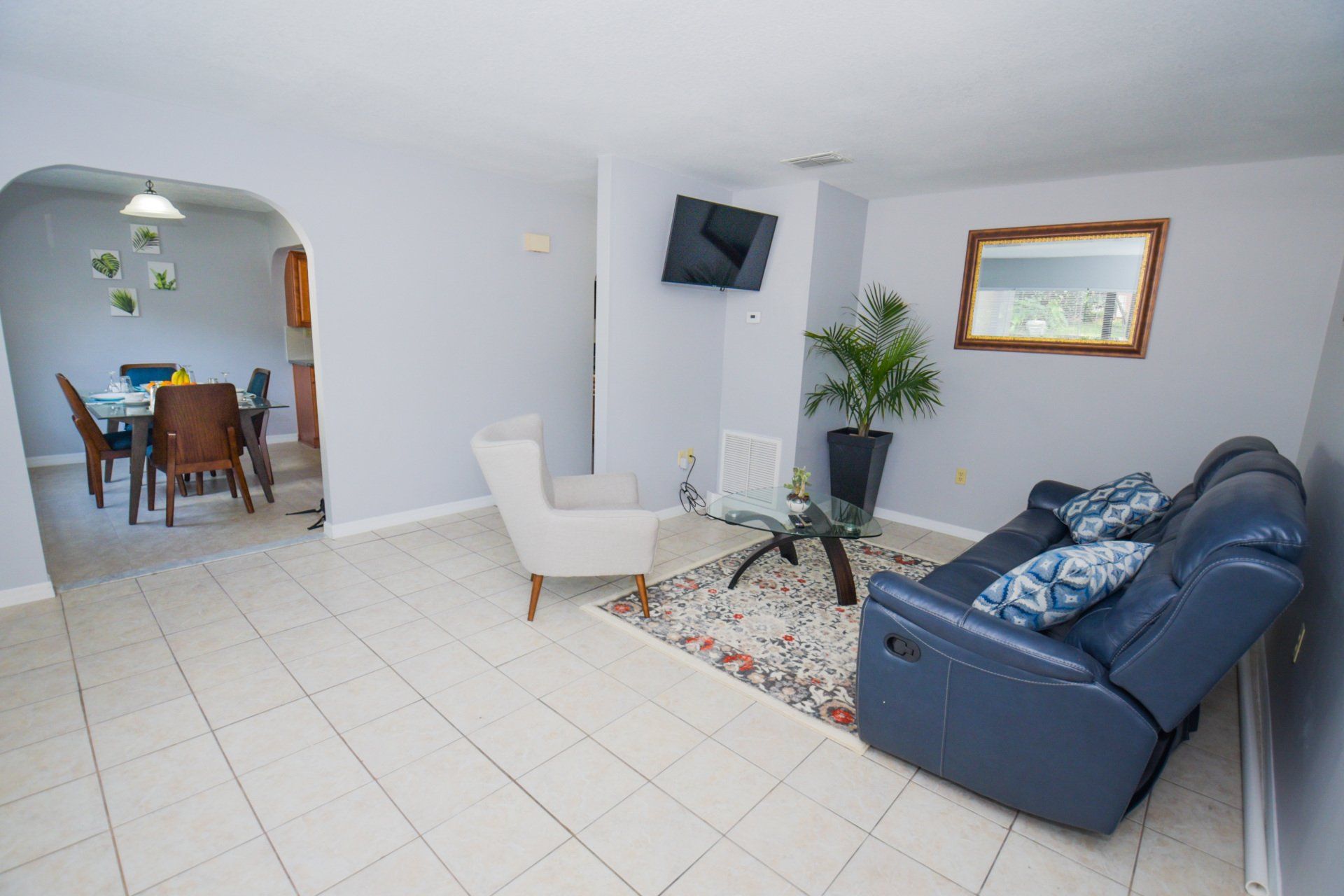 Living room with blue sofa, white chair, glass table, and dining area visible.