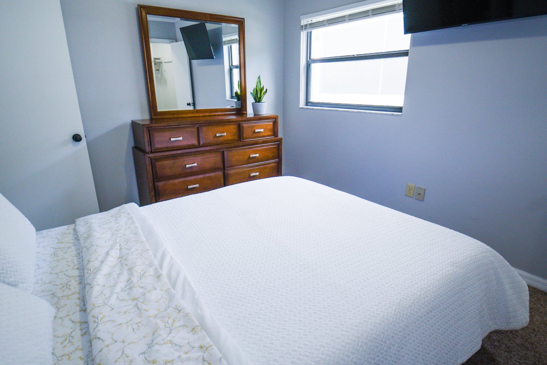 Bedroom with a white bed, wooden dresser, mirror, window, and TV. The walls are light blue.