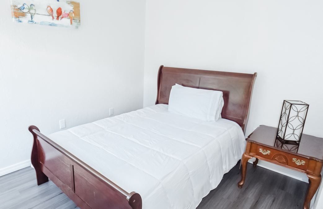 Bedroom with a wooden bed, nightstand, artwork, and white bedding against a light gray wall.