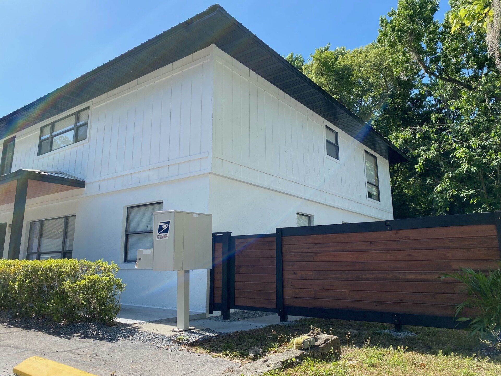 White two-story building with dark trim. A mailbox and wooden fence are in front. Sunny, outdoor setting.