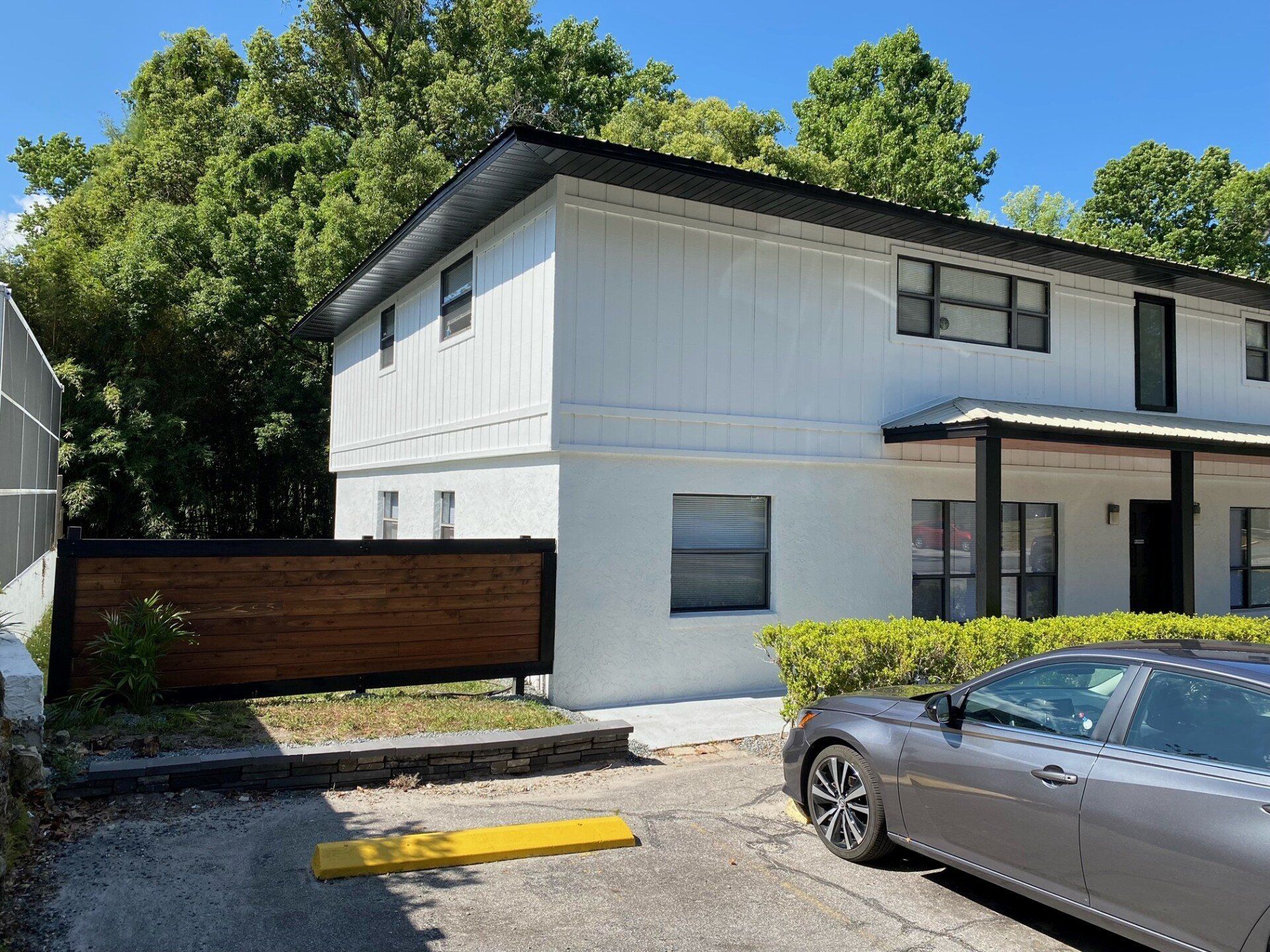Two-story white building with a black roof, wooden fence, and parked car on a sunny day.