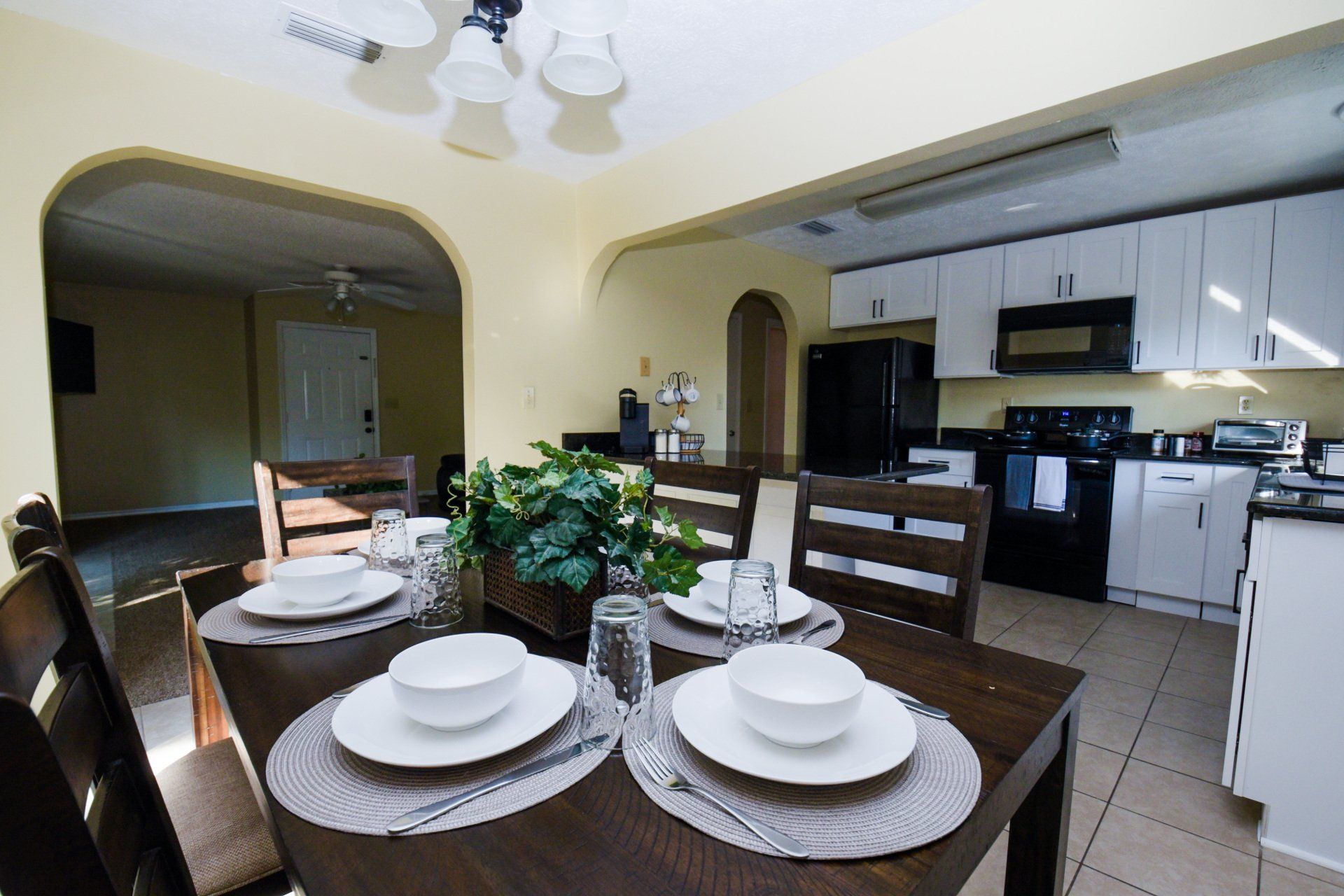 Dining room with wooden table, place settings, and connected kitchen with black appliances.