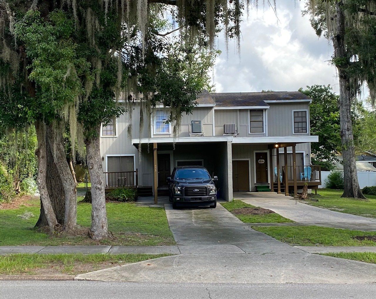 Two-story gray duplex with a black car parked in the driveway. Trees frame the building.