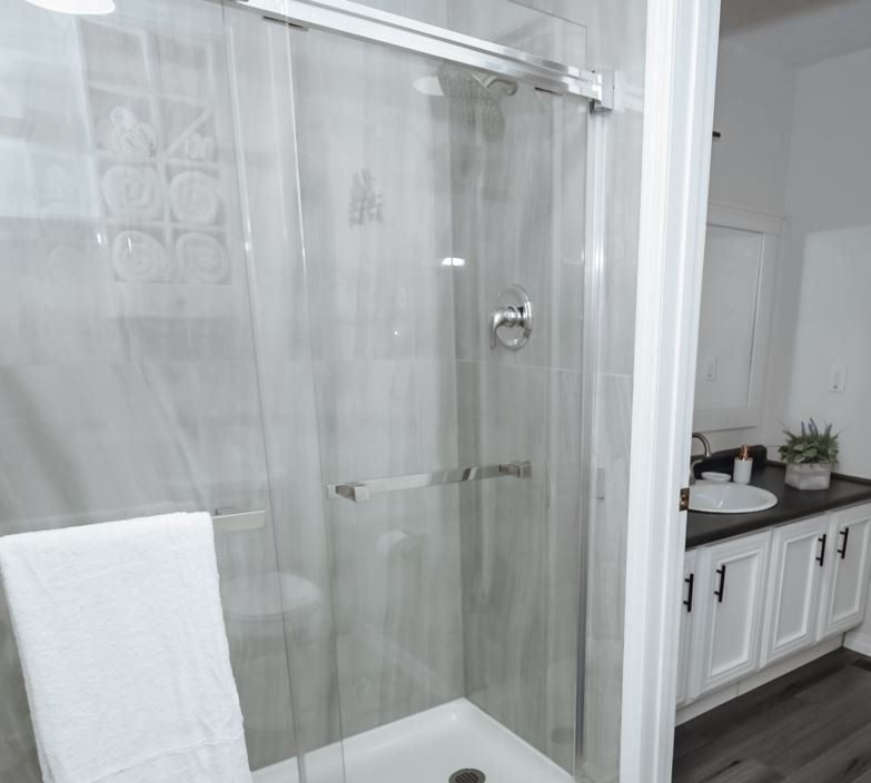 Shower with sliding glass door, white towel, gray tiled walls, and white vanity in bathroom.