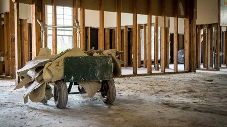 Inside a gutted building; a weathered cart holds debris. Wooden studs frame the rooms; daylight enters through a window.