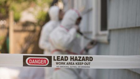 Lead hazard warning tape in front of workers in protective suits removing paint from a building.
