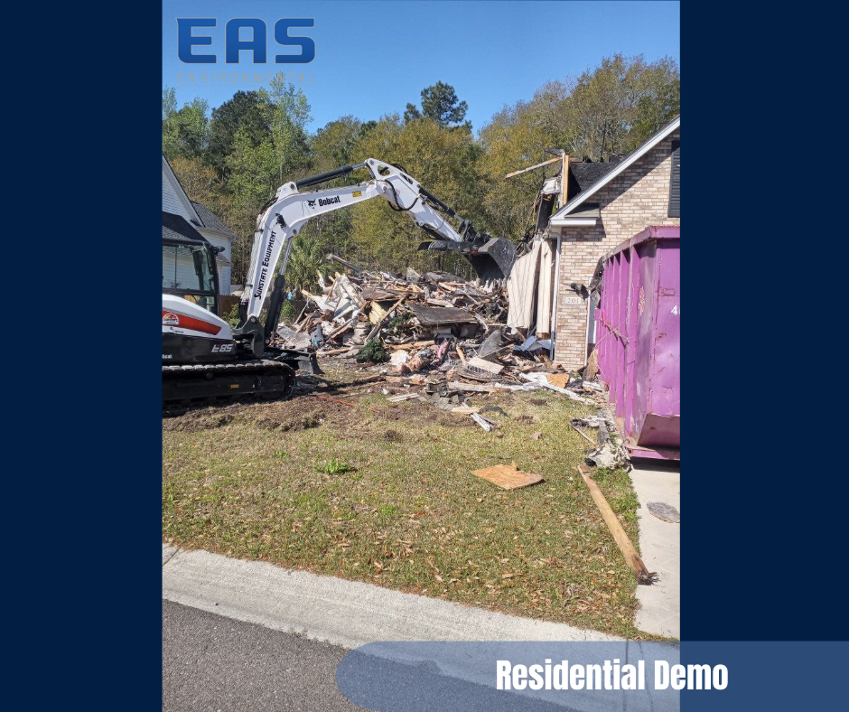A small excavator demolishes a residential house, with a purple dumpster nearby.