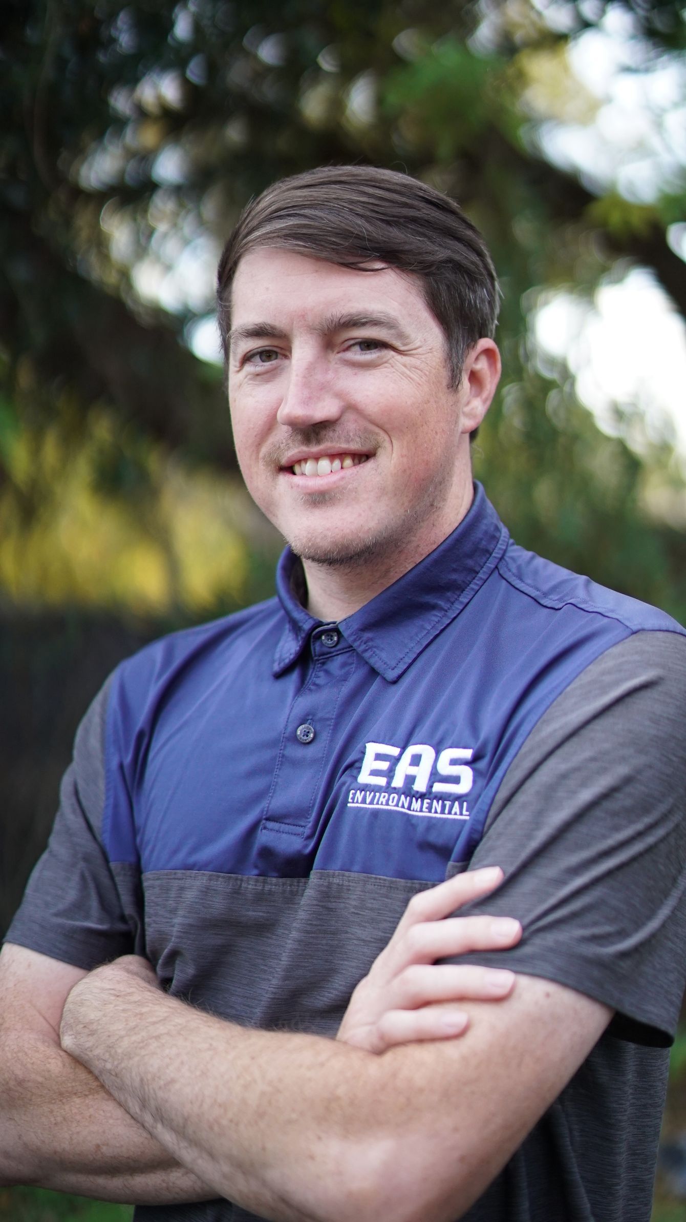 Man with arms crossed, wearing blue and gray polo shirt with 