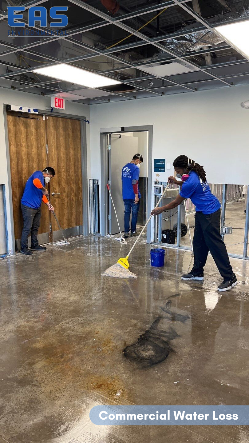 Three people mopping a flooded commercial space. Wet concrete floor, blue shirts, emergency exit sign, and a logo.